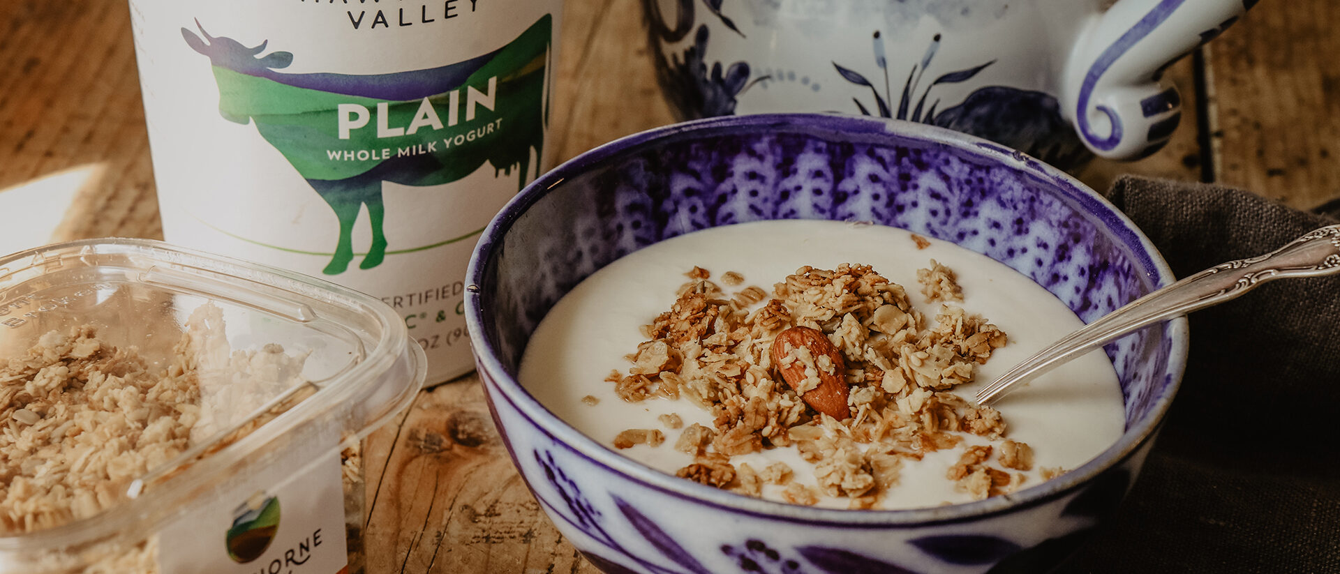 Hawthorne Valley plain yogurt container in the background with a blue and white bowl filled with yogurt and topped with Hawthorne Valley honey almond granola a package of which can be seen in the lower left hand corner