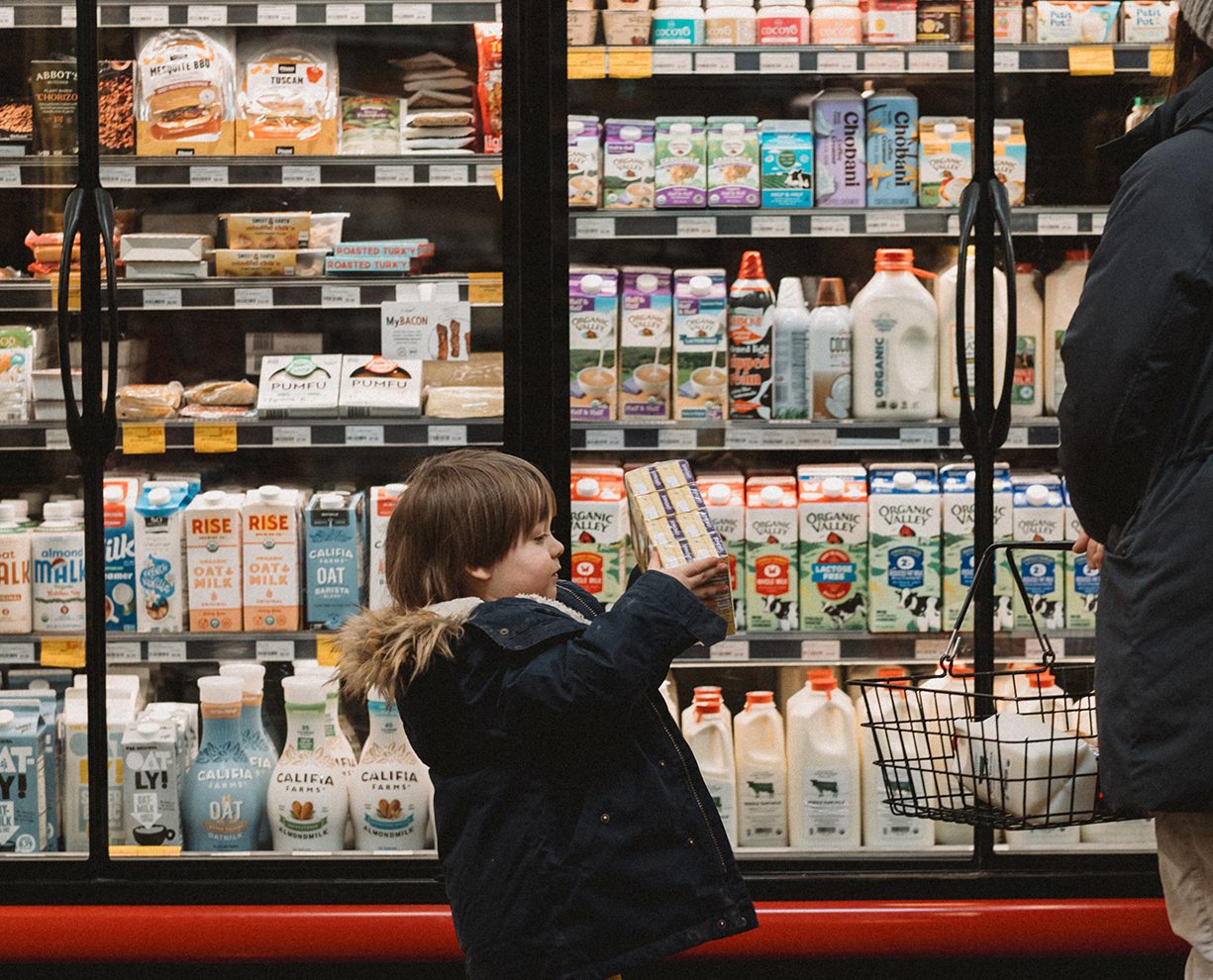 young child bringing juice boxes over to his mother in front of dairy fridges