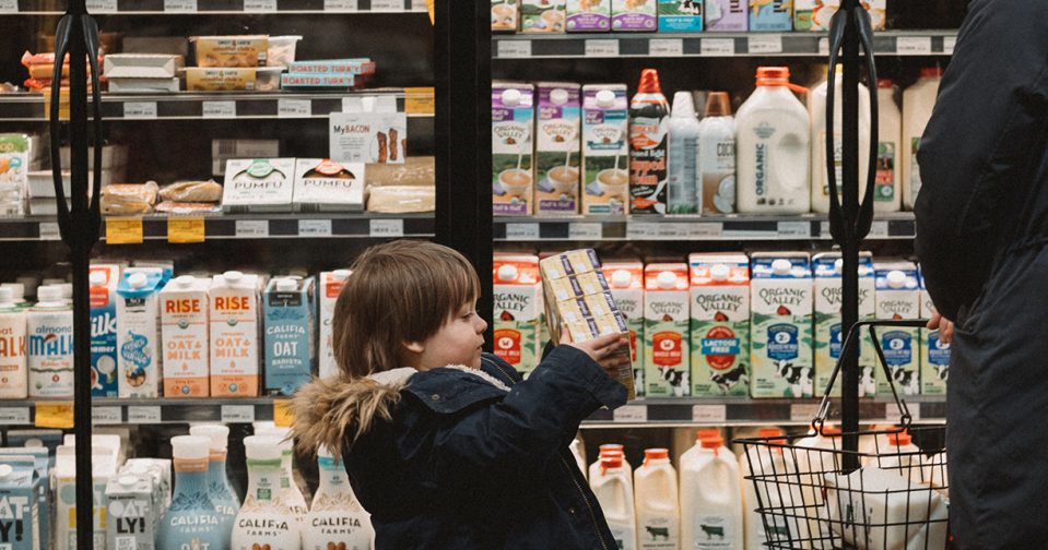 young child bringing juice boxes over to his mother in front of dairy fridges