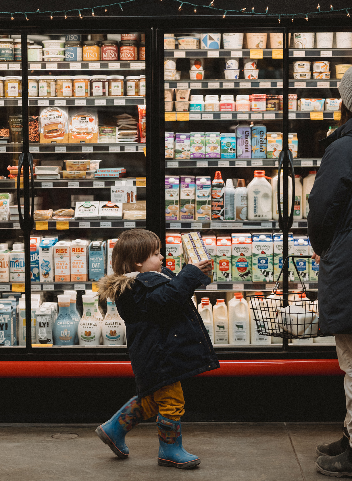 young child bringing juice boxes over to his mother in front of dairy fridges