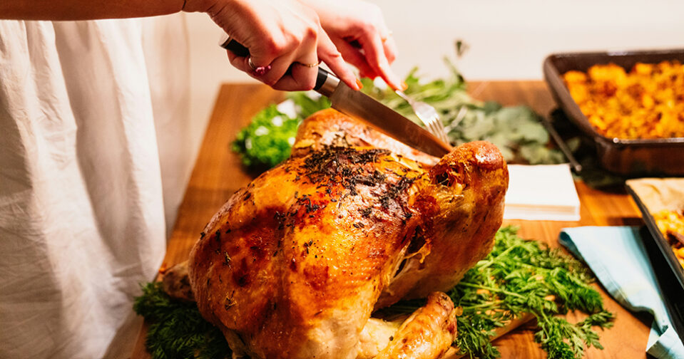 cooked turkey on a cutting board with greenery underneath; set upon a wooden countertop with blurred out casserole dish in background; a woman's hands holding a knife aimed at carving the turkey enter the frame from the top left